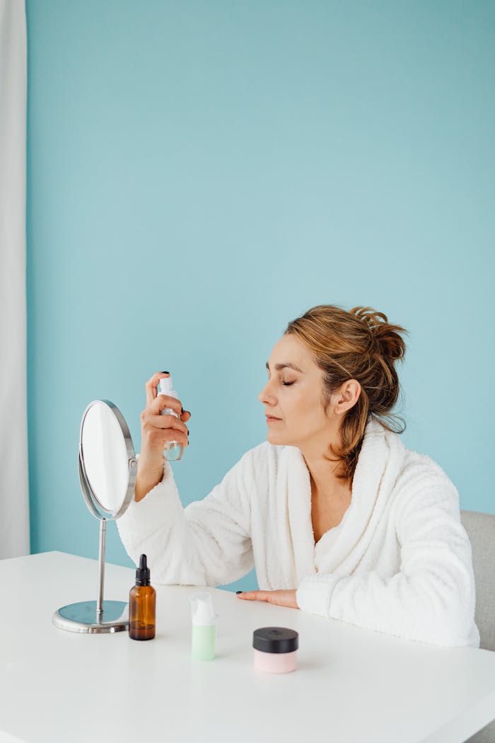 Woman in white bathrobe applying skincare in front of a mirror with closed eyes.